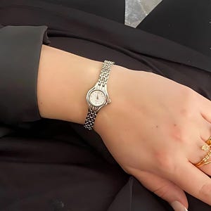 May include: A silver wristwatch with a small, round face and a linked band. The watch is on a wrist, and the person is wearing gold rings. The watch face is white with black numerals.