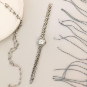 May include: A silver-toned wristwatch with a rectangular face and a linked band. The watch has a white dial with small, delicate hands and markers. A silver beaded necklace and blue threads are also visible.