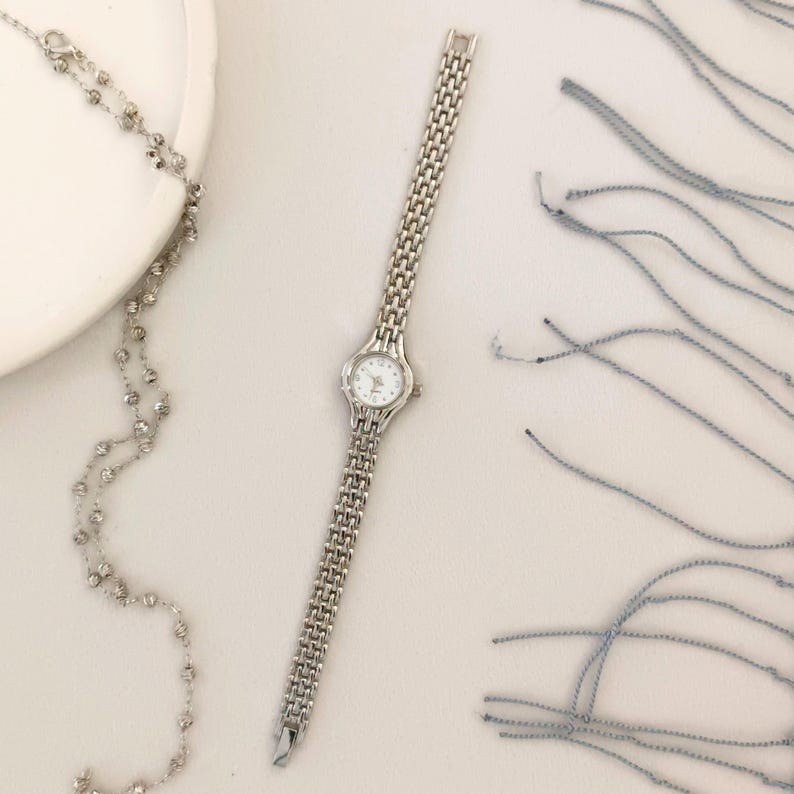 May include: A silver-toned wristwatch with a rectangular face and a linked band. The watch has a white dial with small, delicate hands and markers. A silver beaded necklace and blue threads are also visible.