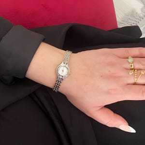 May include: A silver-toned wristwatch with a small, round white face and a linked band is displayed. The watch is worn on a wrist, with a black sleeve in the background. The watch band is approximately 0.5 inches wide.