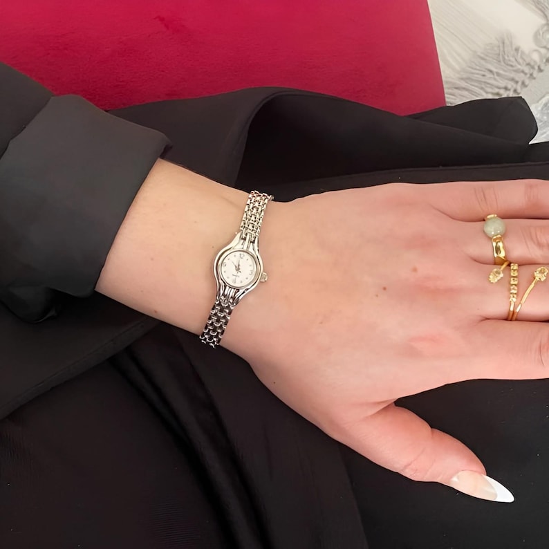 May include: A silver-toned wristwatch with a small, round white face and a linked band is displayed. The watch is worn on a wrist, with a black sleeve in the background. The watch band is approximately 0.5 inches wide.