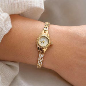 May include: A gold-tone analog wristwatch featuring a small, round white dial with gold-tone hour markers and hands. The watch has a slender, polished gold-tone metal link bracelet with a secure clasp, creating a refined and classic appearance.