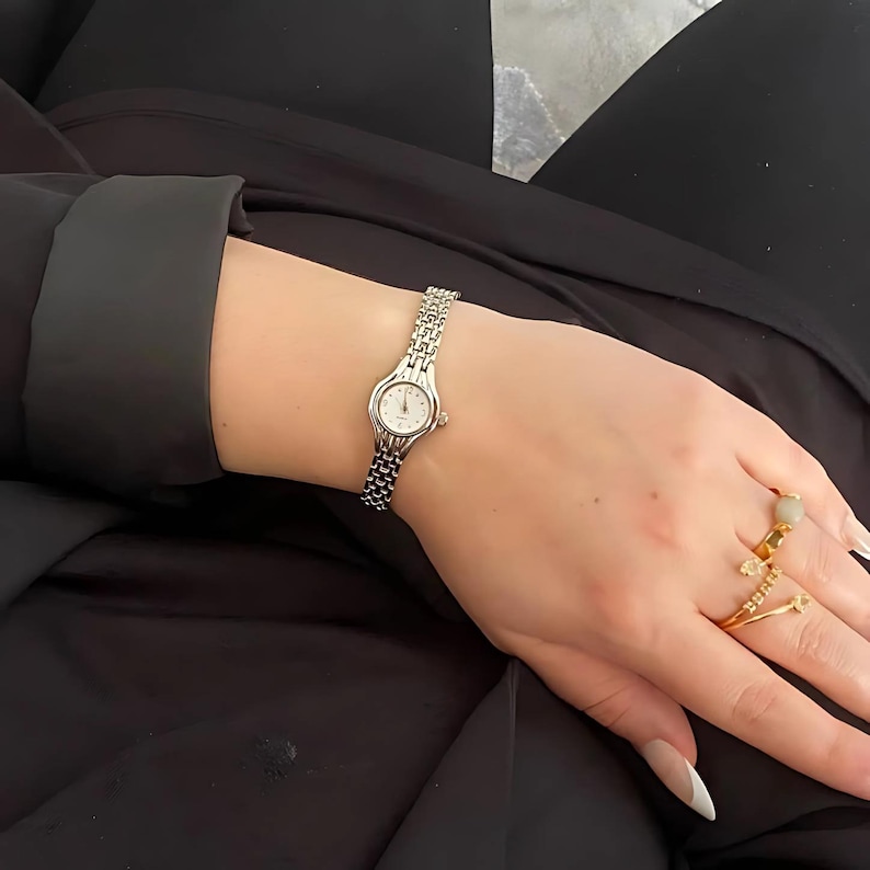 May include: A silver wristwatch with a small, round face and a linked band. The watch is on a wrist, and the person is wearing gold rings. The watch face is white with black numerals.