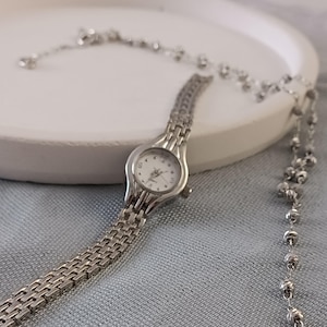 May include: A silver-toned wristwatch with a white face and a linked band. The watch is displayed with a matching silver beaded necklace on a white surface. The background is a light blue fabric.