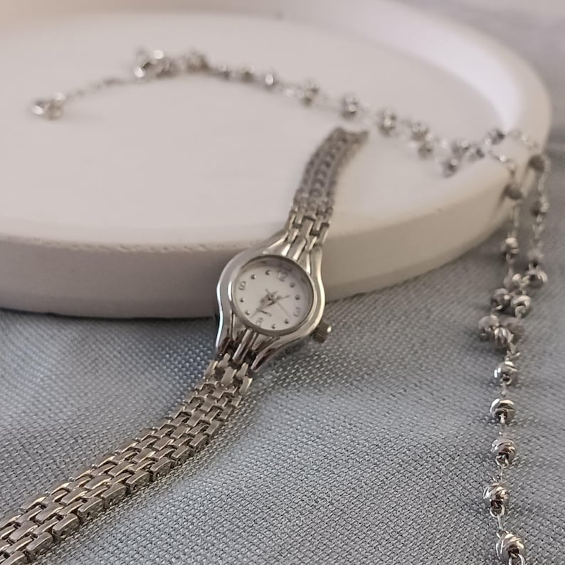 May include: A silver-toned wristwatch with a white face and a linked band. The watch is displayed with a matching silver beaded necklace on a white surface. The background is a light blue fabric.