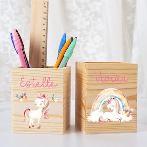 Puede incluir: Dos portalápices de madera con diseños de unicornios. Uno tiene el nombre "Estelle" y un unicornio con mariposas. El otro tiene el nombre "Vivian" y un unicornio con un arcoíris. Bolígrafos y una regla en el interior.