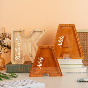 Può includere: Tre salvadanai in legno a forma di lettera. Uno è una "K" in legno chiaro con "Kalinda" in scrittura bianca. Due "A" sono in legno più scuro, ciascuno con "Adele" in scrittura bianca. I salvadanai sono su una superficie bianca.