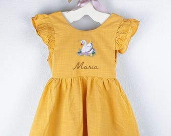 May include: Mustard yellow dress with ruffled sleeves. The dress features an embroidered swan design with floral accents and the name "Maria" in script. The dress is hanging on a white hanger with a decorative swan and pink ribbon.