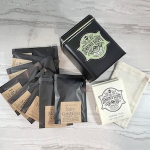 May include: A black metal tin with a green and black label that reads "Dem Bones Shoppe" and "Loose Leaf Tea Sampler". The tin is filled with eight individual tea bags wrapped in black paper. The tea bags are labeled with names like "Fairy GothMother" and "Witchy Green". The tea bags are accompanied by a white muslin bag and a white card with the same label as the tin.