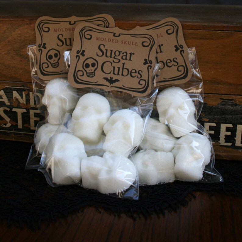 Moody gothic photo of 3 bags of skull shaped sugar cubes propped against a vintage wood box. Label reads Moulded Skull Sugar Cubes.