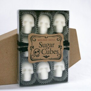 Box of 9 skull shaped sugar cubes rests on kraft coffin box, each skull is nested in a black candy cup, black ribbon around box with kraft label: Sugar Cube Skulls,  all on white background