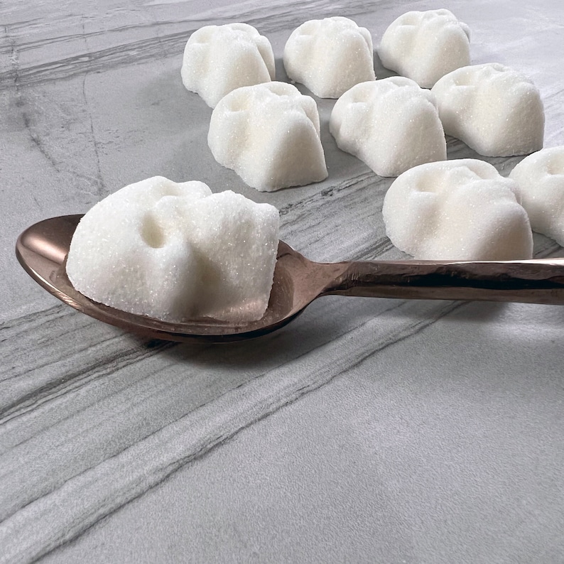 Close up view of a skull shaped sugar cubes on a gold spoon. More sugar cubes are in the background on a grey tile surface.