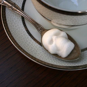 Cropped view of white tea cup and saucer with a silver rim sitting on a black background. A vintage silver spoon rests on the saucer with skull shaped sugar cube in it.