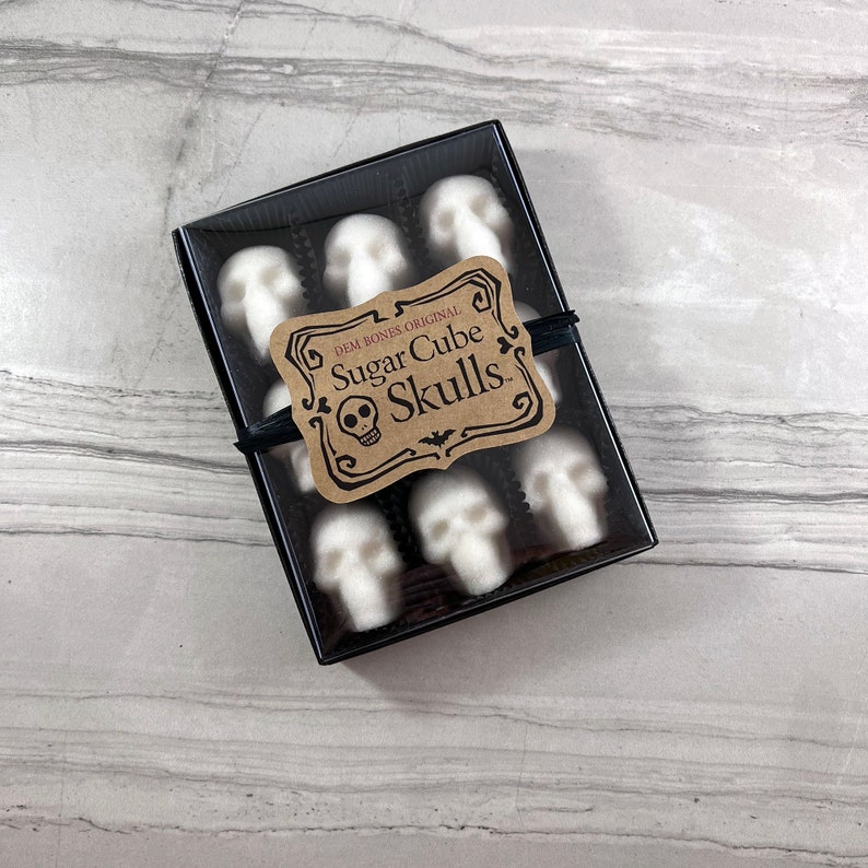 Tile background with box of skull shaped sugar cubes, each sugar cube sits in a candy cup in a grid, Kraft labels read: DemBones Original, Sugar Cube Skulls with skull and bat artwork