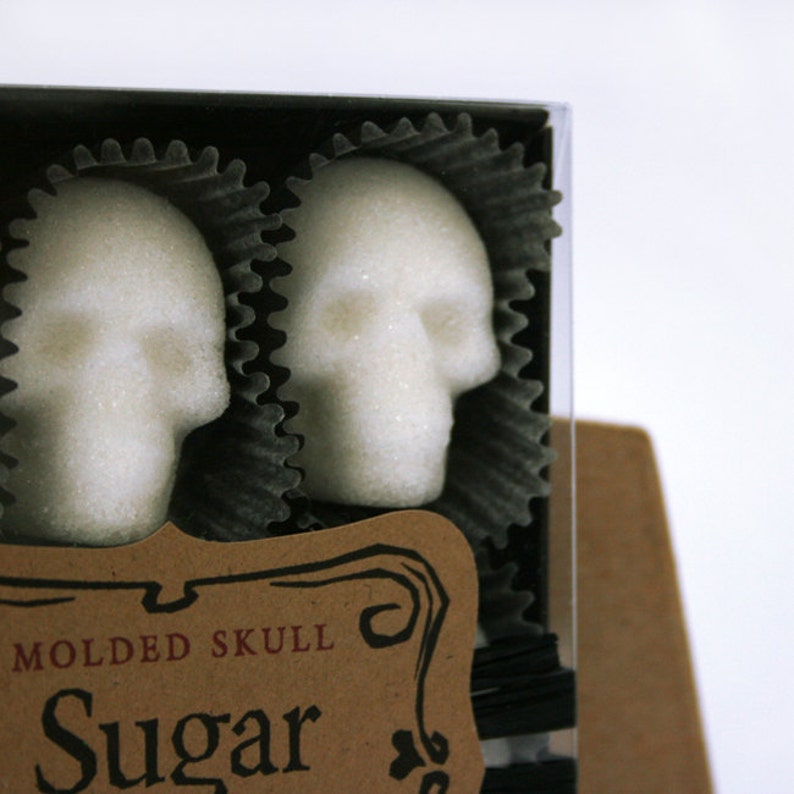Close up of corner of a box featuring skull shaped sugar cubes in black candy cups. Corner of kraft label reads Sugar. White background with no props