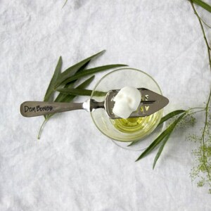 Top view of absinthe glass with skull sugar cube on absinthe spoon balanced on glass rim, White table cloth with sprig of greenery on left top