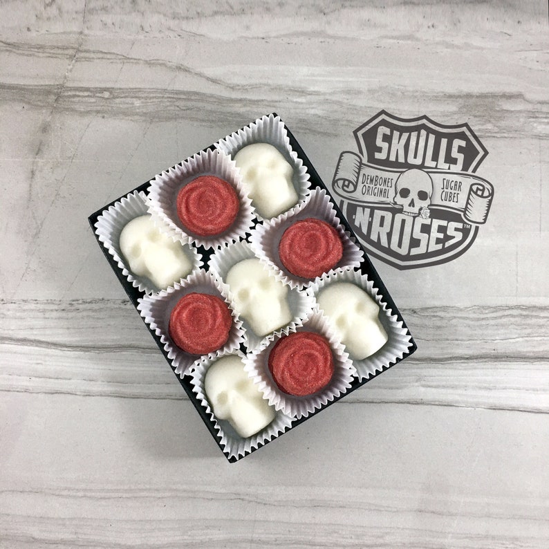 Tile background with open box of skull shaped sugar cubes & red rose shaped sugar cubes, each sugar cube sits in a white candy cup in a grid, Rubber stamp logo over image reads: Skulls N Roses, DemBones Original, Sugar Cubes