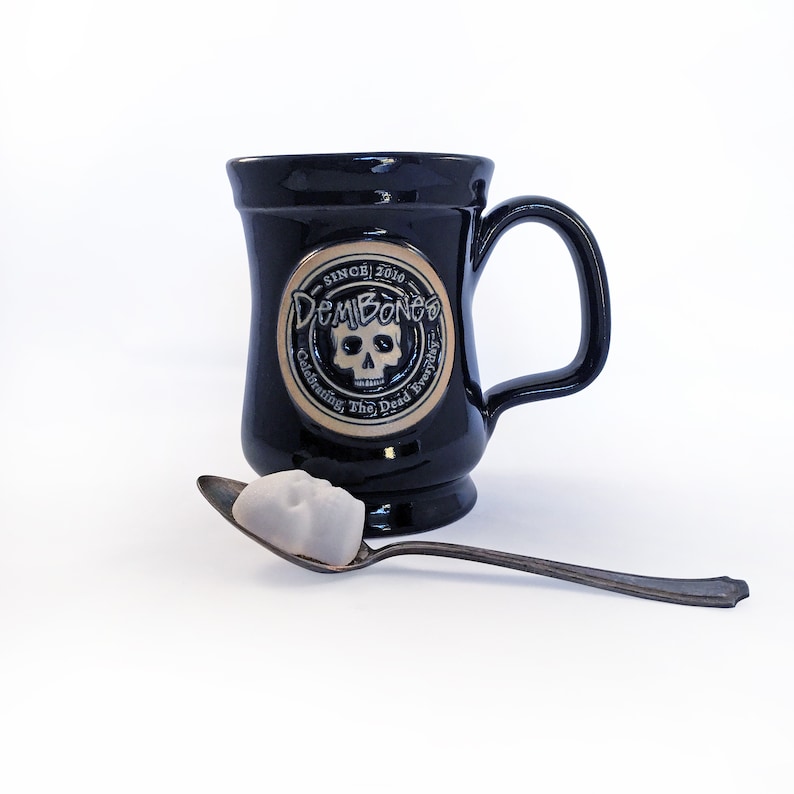 Black ceramic mug with DemBones round logo, a single skull shaped sugar  cube sits on a vintage silver spoon in foreground, Wording reads DemBones Since 2010, celebrating the dead everyday