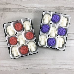 Tile background with 2 open boxes of skull shaped and rose shaped sugar cubes, each sugar cube sits in a white candy cup in a grid, Right side has red roses left side has purple roses,