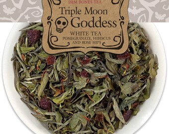 Op de afbeelding: Een witte kom gevuld met losse bladthee. De thee is een mix van witte thee, granaatappel, hibiscus en rozenbottels. Een bruin label met de tekst "DEM BONES TEA Triple Moon Goddess WHITE TEA POMEGRANATE, HIBISCUS AND ROSE HIPS" bevindt zich in de linkerbovenhoek van de afbeelding.