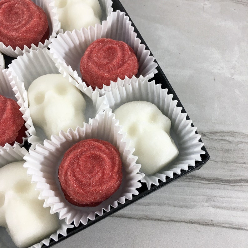 Close up of a box of skull shaped sugar cubes and red rose shaped sugar cubes, each sugar cube sits in a white candy cup in a grid, tile background