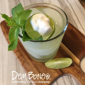 overhead view of Sugar Skull Mojito, Mojito cocktail with skull sugar cubes on lime slice, green mint in glass on wood cutting board and tile counter, DemBones celebrating the dead everyday logo in left corner