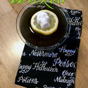 Wood table overhead view of a black cocktail with a slice of lemon floating in it and a skull shaped sugar cube on top. Glass sits on a black Halloween dish towel. Green text above photo reads Wicked and DemBones logo is in bottom left corner.