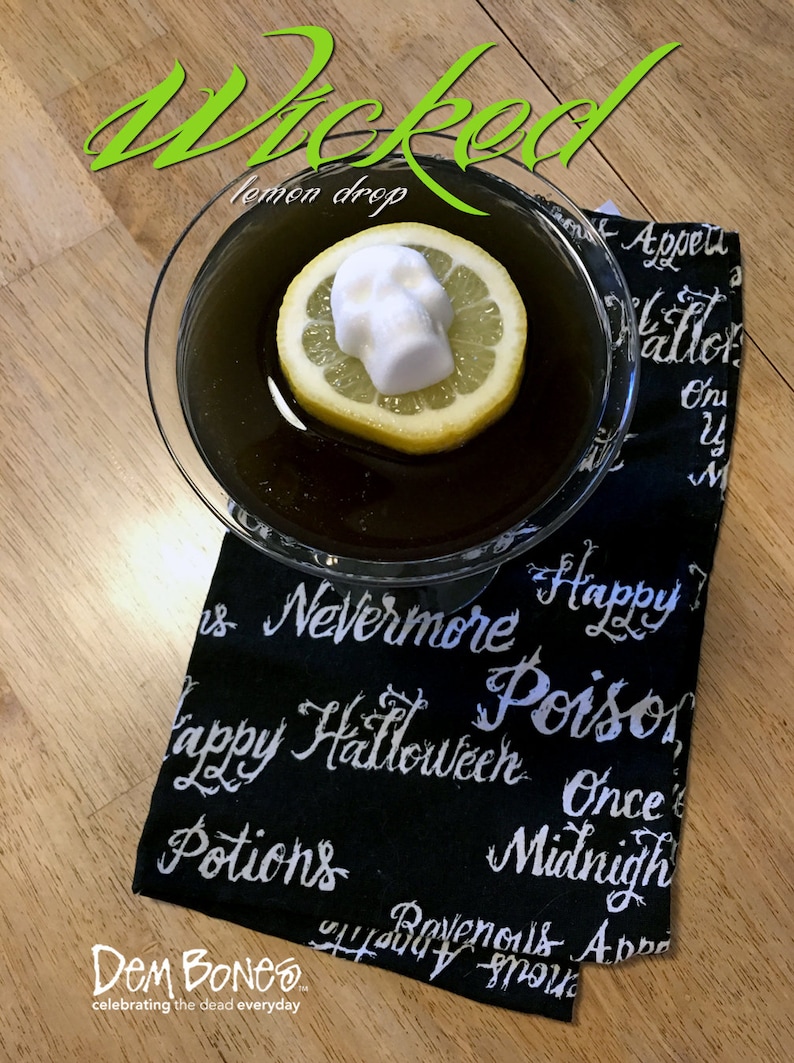 Wood table overhead view of a black cocktail with a slice of lemon floating in it and a skull shaped sugar cube on top. Glass sits on a black Halloween dish towel. Green text above photo reads Wicked and DemBones logo is in bottom left corner.