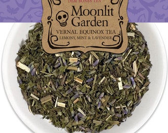 Op de afbeelding: Een witte kom gevuld met losse bladthee. De thee is een mix van groene bladeren, lavendelknoppen en kleine stukjes citroengras. Een bruin papieren etiket met de tekst "Moonlit Garden Vernal Equinox Tea Lemony, Mint & Lavender" is zichtbaar op de achtergrond.