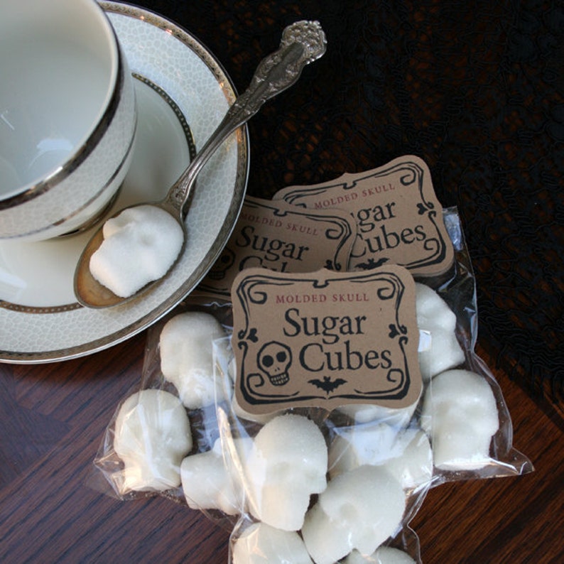 Moody gothic overhead view, DemBones Sugar Cube Skulls in bags scattered on dark wood table, tea cup and saucer with silver spoon and skull sugar cube on edge,