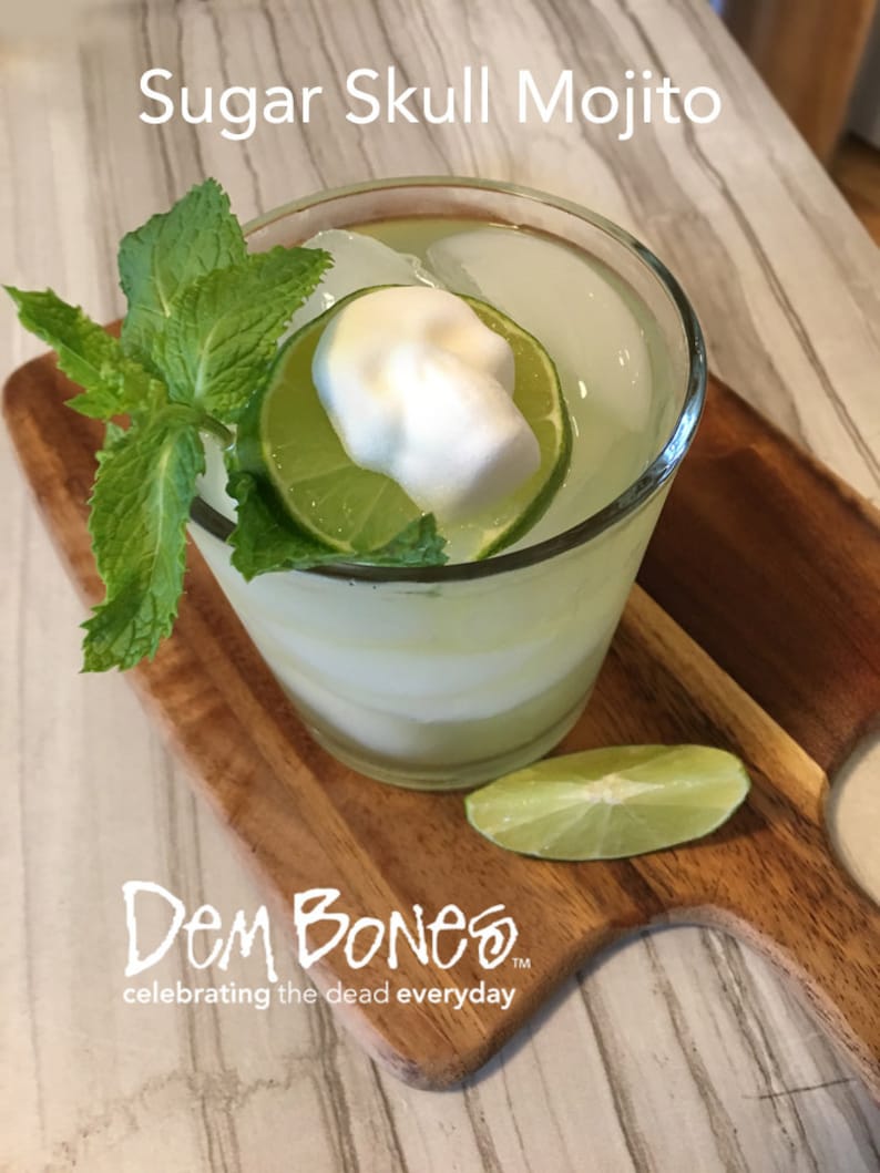 overhead view of Sugar Skull Mojito, Mojito cocktail with skull sugar cubes on lime slice, green mint in glass on wood cutting board and tile counter, DemBones celebrating the dead everyday logo in left corner