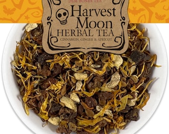 Op de afbeelding: Een witte kom gevuld met een mengsel van losse kruidenthee. De thee is een mix van gedroogde bloemen, kruiden en specerijen. Het etiket op de kom luidt "Harvest Moon Herbal Tea Cinnamon, Ginger & Apricot."
