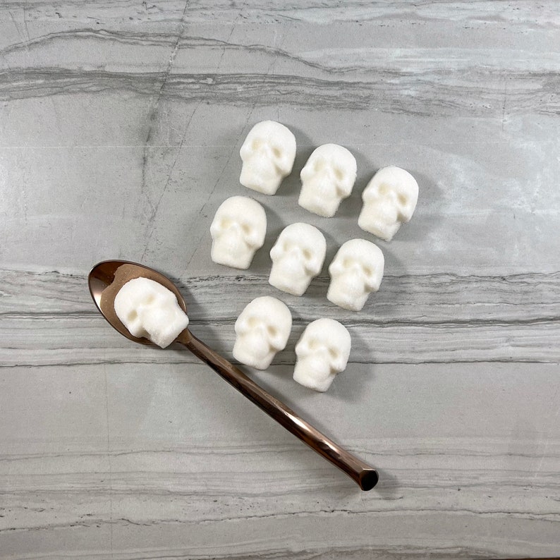 Overhead view of eight skull shaped sugar cubes lined up in a grid on grey tile surface. One skull sits in a gold spoon at an angle  in front of the rest of the sugar cubes.