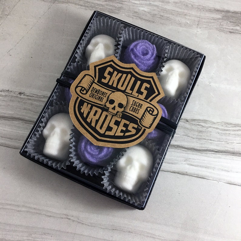 Tile background with box of skull shaped sugar cubes and purple rose shaped sugar cubes, each sugar cube sits in a candy cup in a grid, Kraft labels read: Skulls N Roses, DemBones Original, Sugar Cubes
