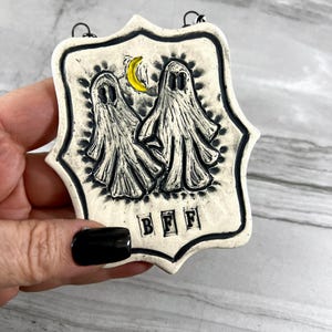 May include: White ceramic ornament with two black and white ghosts and a yellow crescent moon. The ornament has a black border and the word "BFF" written in black letters.