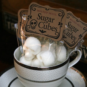 DemBones Skull shaped Sugar Cubes, Sugar cubes in bags with kraft labels sitting in a white tea cup and saucer, darkly gothic and  moody background