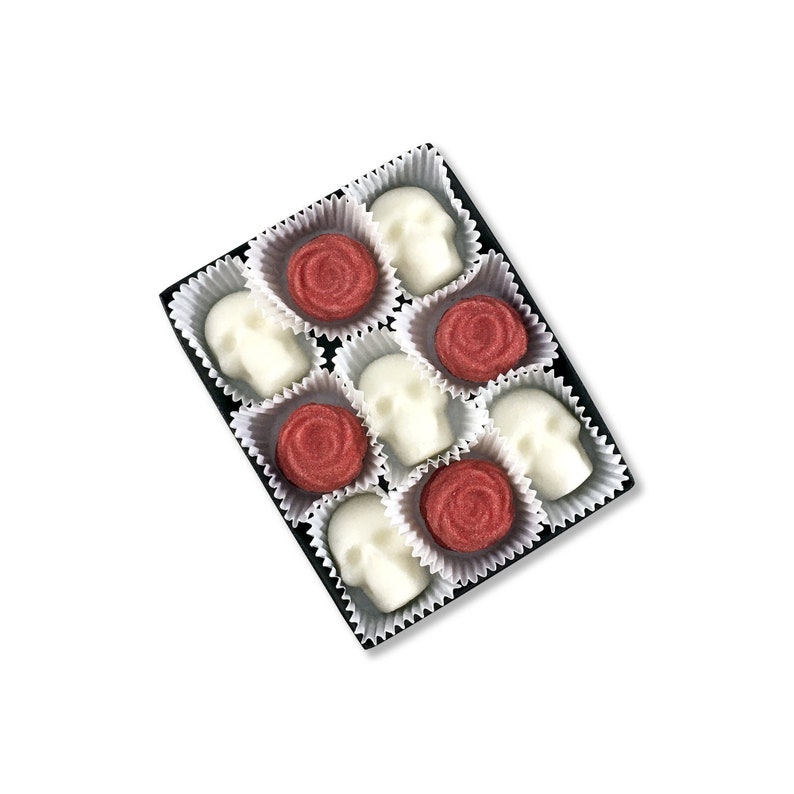 White background with open box of skull shaped sugar cubes & red rose sugar cubes. Each sugar cube sits in a white candy cup in a black box.