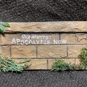May include: A rectangular, brick-like plaque with the text "OUR MOTTO: APOCALYPSE NOW" in white. The plaque is tan with green plant-like embellishments on the edges. The background is a dark, textured surface.