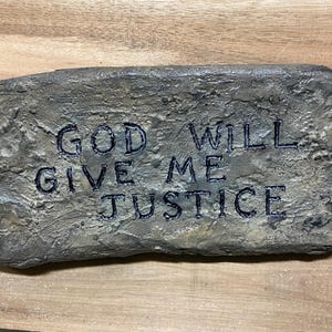 May include: A rectangular, gray stone with the engraved words "GOD WILL GIVE ME JUSTICE" in dark blue. The stone has a rough, textured surface and is set against a wooden background.