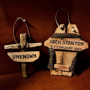 May include: Two wooden ornaments, one shaped like a cross with the word "UNKNOWN" written on it, and the other shaped like a small wooden sled with the text "ARCH STANTON 3 FEBRUARY 1862" written on it.