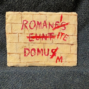May include: A brick wall with the words "ROMANES EUNT DOMUS" written in red paint. The word "EUNT" is crossed out.