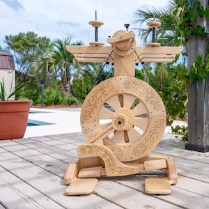 May include: A wooden spinning wheel with a large, floral-decorated wheel and intricate carvings. The wheel has a natural wood finish and is set on a wooden deck outdoors, with a pool and greenery in the background.