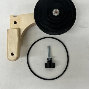 May include: A disassembled wooden device with a black wheel, a black rubber ring, a bolt, and a black knob. The wooden component is light-colored, and the wheel has concentric circles. The bolt and knob are metallic and black.