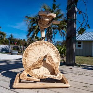 May include: A wooden spinning wheel with floral engravings. The wheel features a large, circular design with spokes and decorative floral patterns around the rim and on the arms. The natural wood color contrasts against the blue sky and outdoor setting.