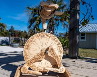 May include: A wooden spinning wheel with floral carvings. The wheel features a large, circular design with spokes and decorative floral patterns around the rim. The wheel is made of light-colored wood and is set against a bright blue sky.