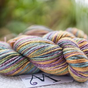 May include: A skein of hand-dyed yarn featuring a variegated color palette of soft lavender, muted sage green, and warm golden yellow. The twisted fiber displays a multi-colored appearance with visible plies. A white tag with a black logo is visible.