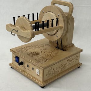 May include: A wooden spinning wheel with a natural finish. The wheel features a handle, a flyer with black pegs, and a base with floral engravings. It has a speed switch and an on/off switch.