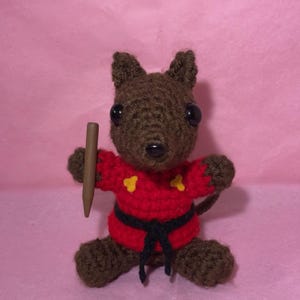 May include: A handmade brown crochet animal with large black eyes, wearing a red karate uniform with yellow accents and a black belt. It holds a brown crayon. The background is pink.