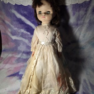 May include: A doll with brown hair and large eyes wearing a long-sleeved, off-white dress. The dress features a high neckline with vertical lace trim, a satin ribbon waistband, and a skirt with eyelet embroidery and a scalloped floral hem.