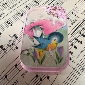 May include: A small, rectangular, pink trinket box with a painted scene of a blue bird, pink mushroom, and purple and yellow flowers. The box is on a sheet of vintage sheet music.